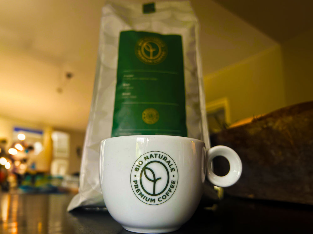 Een kopje van Bio Naturale Premium Koffie, met de groene zak biologische koffiebonen op de achtergrond. De koffie, gemaakt van 100% Arabica bonen, biedt een rijke smaak met fruitige en karamelsmaken. Verkrijgbaar bij CaféHuis voor de liefhebber van biologische, premium koffie.