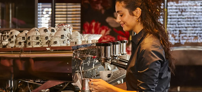 Een barista bereidt koffie met Brizio kopjes op de espressomachine in een sfeervol café. Brizio staat voor authentieke Italiaanse koffiekwaliteit, geliefd in de horeca en bij koffieliefhebbers wereldwijd.