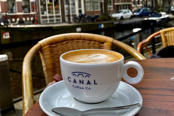 Canal Coffee Co. cappuccino geserveerd in een wit kopje op een terras met uitzicht op de iconische Amsterdamse grachten. Perfecte combinatie van koffiebeleving en Nederlandse charme.