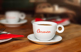 Carmen koffiekopje met rood logo, geserveerd op een houten tafel met rode accenten, onderdeel van het exclusieve assortiment van CaféHuis. Een premium koffiebeleving met een klassieke en verfijnde uitstraling.