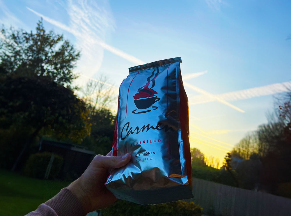 Een zak Carmen Superieur koffiebonen vastgehouden tegen een prachtige ochtendhemel. De perfecte start van de dag met premium koffie vol smaak en karakter.