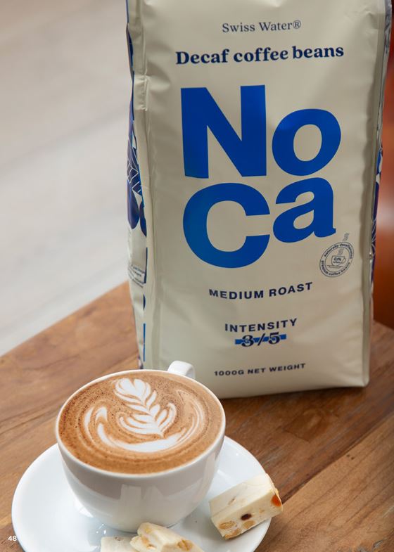 NoCa Decaf Coffee Beans – 1000g cafeïnevrije koffiebonen, medium roast met intensiteit 3/5, gepresenteerd naast een kopje romige cappuccino met latte art en een zoete lekkernij. Swiss Water® proces voor een verfijnde cafeïnevrije koffie-ervaring.