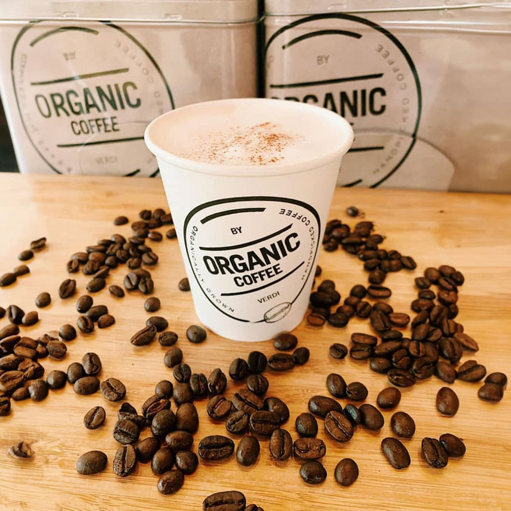 Organic Coffee van CaféHuis – biologisch geteelde koffie gepresenteerd in een stijlvolle to-go beker, omringd door vers geroosterde koffiebonen. Perfect voor een duurzame en luxe koffie-ervaring onderweg